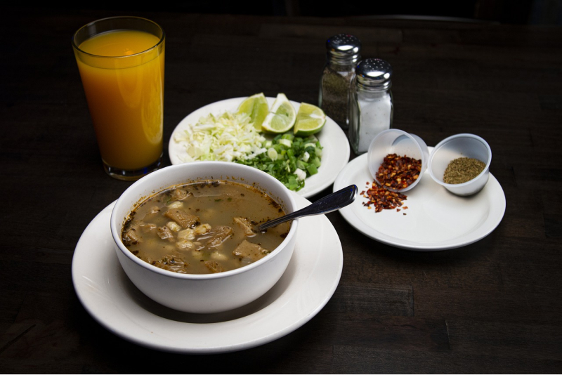Un bol de menudo, mâncare mexicană tradițională, lângă un pahar de suc și farfurii cu salată și condimente